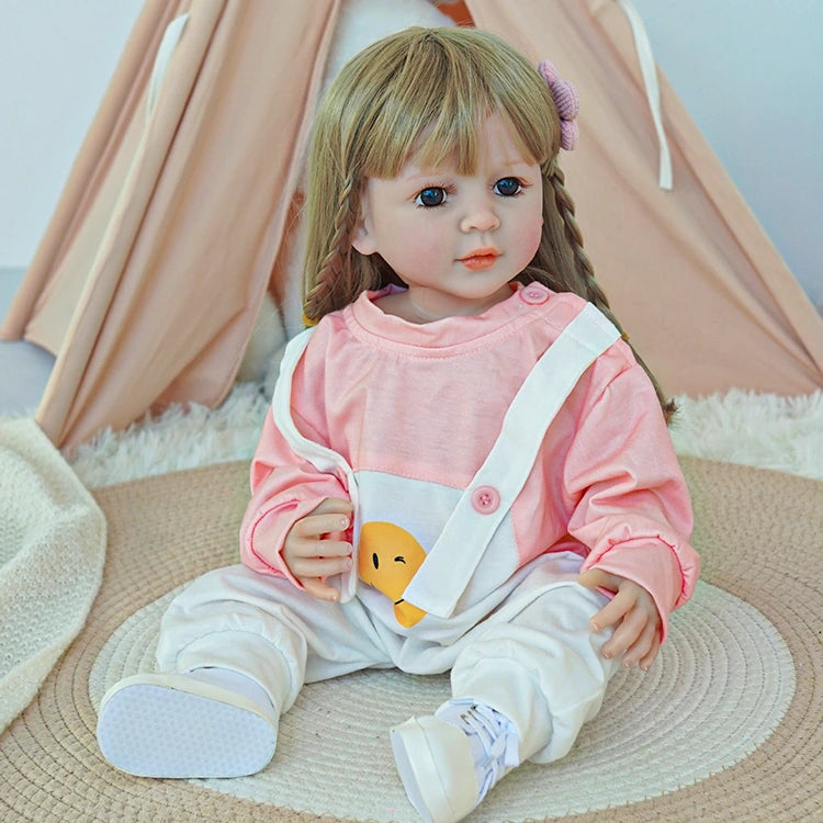 Reborn toddler doll sitting on a soft rug, wearing a pink top and white overalls, looking ahead with a gentle expression.