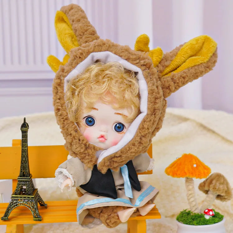 An adorable doll with blue eyes and a cozy bunny hat, situated among playful miniatures including a tiny Eiffel Tower.