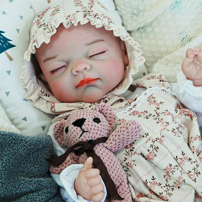 The reborn baby doll in a calm pose, dressed in a floral outfit with a matching bonnet, holding a knitted teddy bear close.