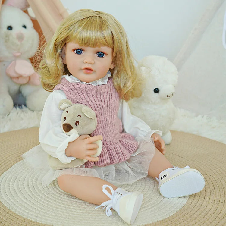 Close-up of reborn baby doll with blonde hair in a pink knit dress holding a teddy bear.