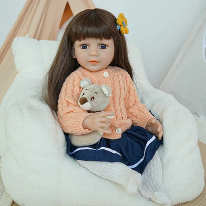 Reborn baby doll in peach sweater and navy skirt, holding a teddy bear, sitting in a white chair.