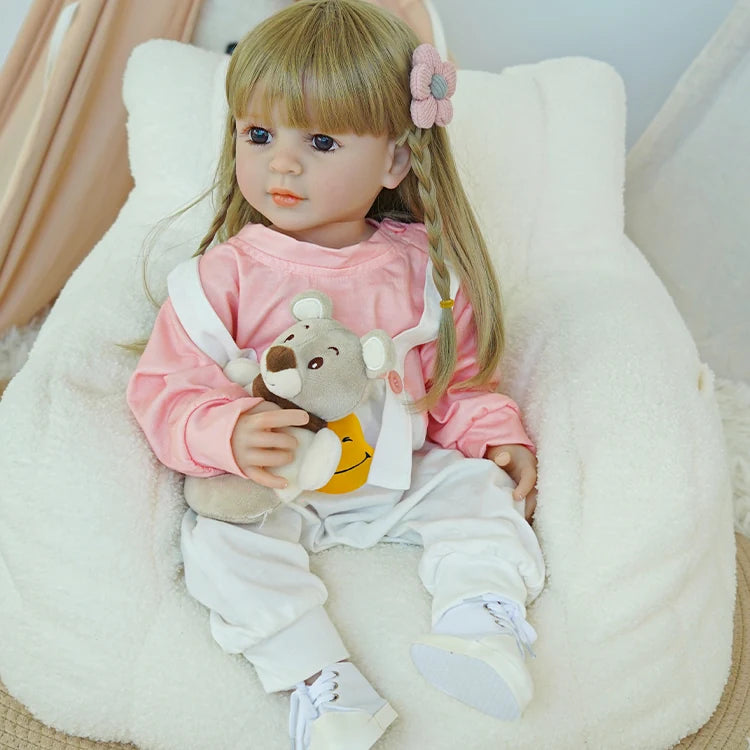 Reborn baby doll in pink shirt and white overalls, sitting on a rug, holding a teddy bear.