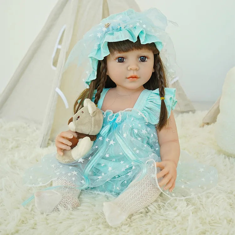 Reborn toddler doll in a blue starry dress and hat, holding a teddy bear, seated on a fluffy white carpet.