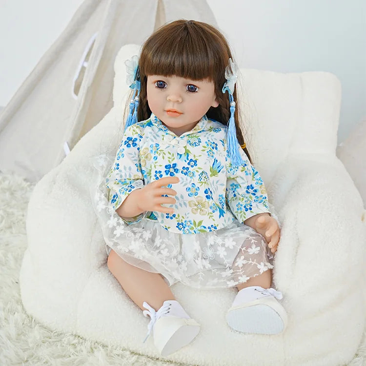 Toddler reborn doll in a blue floral top, holding a teddy bear, sitting on a white fluffy carpet.