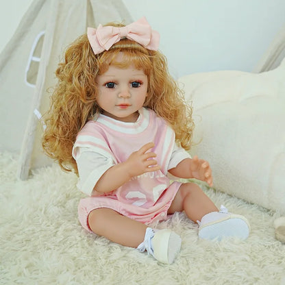 Blonde toddler reborn doll in a pink dress, holding a teddy bear, seated on a fluffy white carpet.