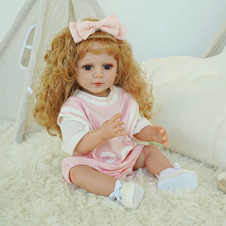 Blonde toddler reborn doll in a pink dress, holding a teddy bear, seated on a fluffy white carpet.