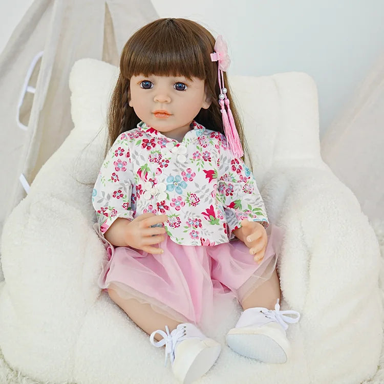 Toddler reborn doll holding a teddy bear, dressed in a floral top and pink skirt, sitting on a cozy white chair.