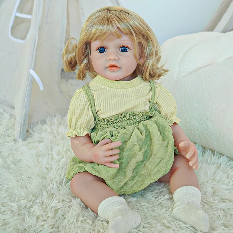 Reborn toddler doll sitting on a rug, wearing a green outfit with a light green top and white socks.