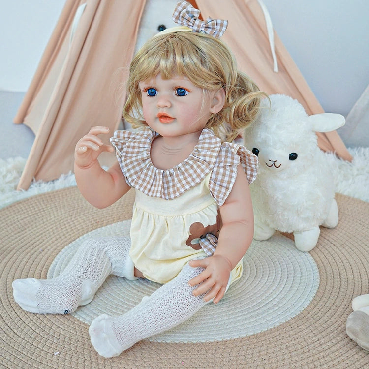  Reborn toddler doll sitting in a plush chair, wearing white overalls with a bunny design, holding a teddy bear.